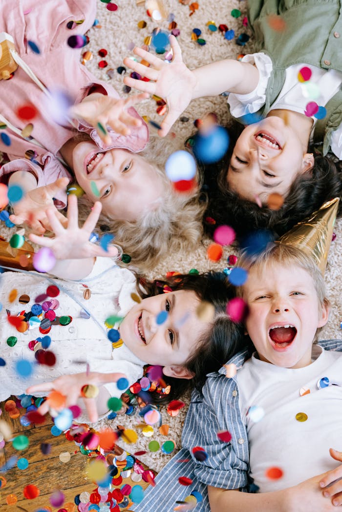 Four cheerful children lying on the floor, playfully surrounded by vibrant confetti.